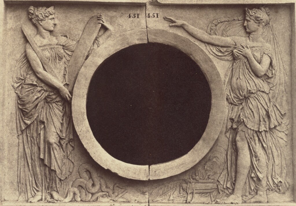 Historic photograph of two classical female allegorical figures positioned around a large circular opening in an architectural relief.
